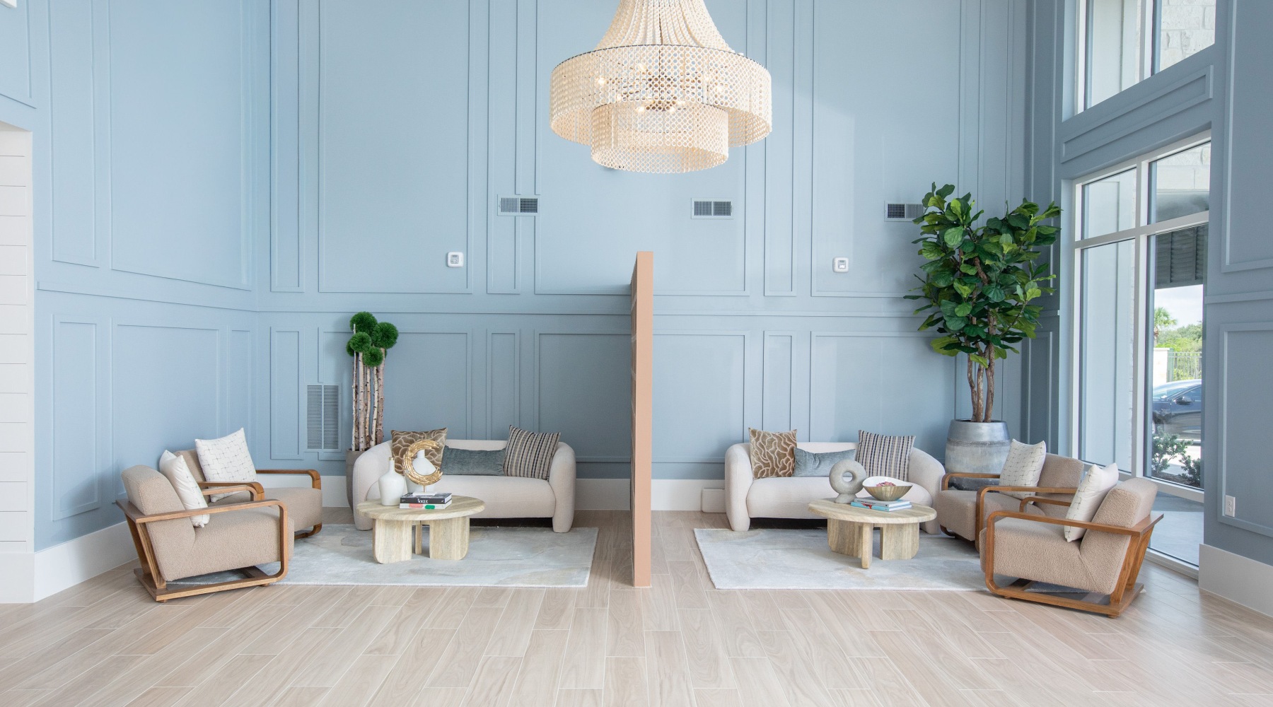 Elegant lounge area with light blue walls, a decorative chandelier, and two seating arrangements divided by a vertical panel. Features include cream-colored sofas, wooden tables, and lush plants.