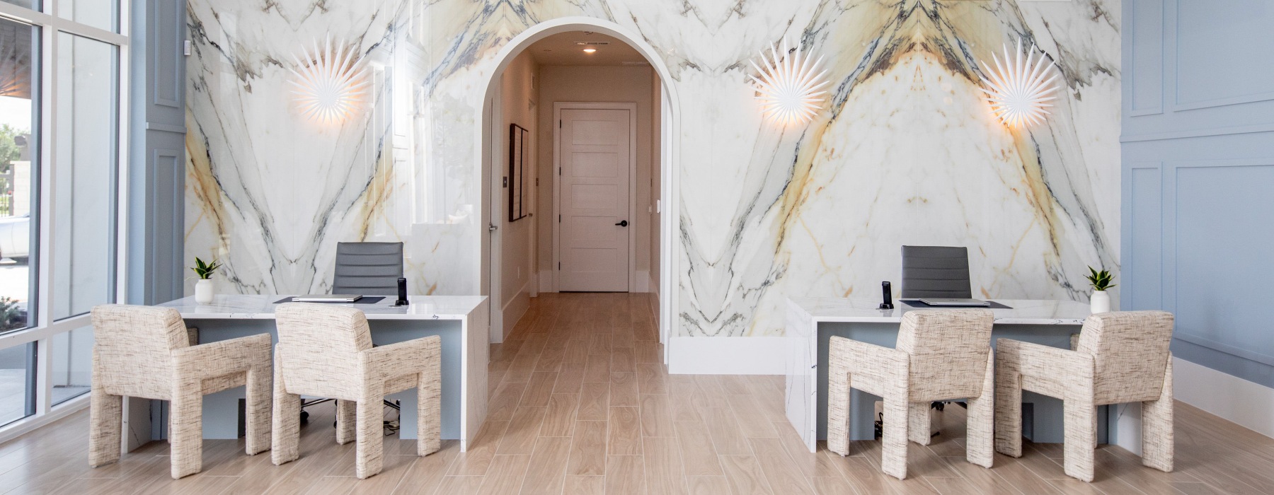 Stylish interior with a large marble wall, two modern desks and chairs, a chandelier, and sunburst light fixtures.