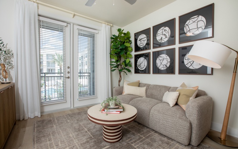 A modern living room with a light gray sofa, striped round coffee table, decorative pillows, and six circular black-and-white artworks on the wall. Large windows with sheer curtains allow natural light and show palm trees and a pool outside.
