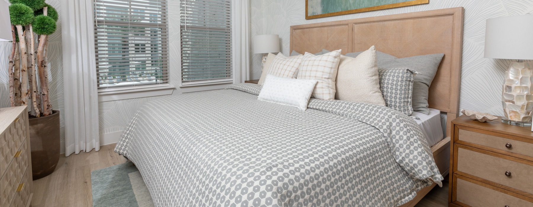 A contemporary bedroom with a king-size bed featuring a patterned duvet, decorative pillows, a textured headboard, and large windows. A nightstand with a lamp and a potted topiary are also visible.