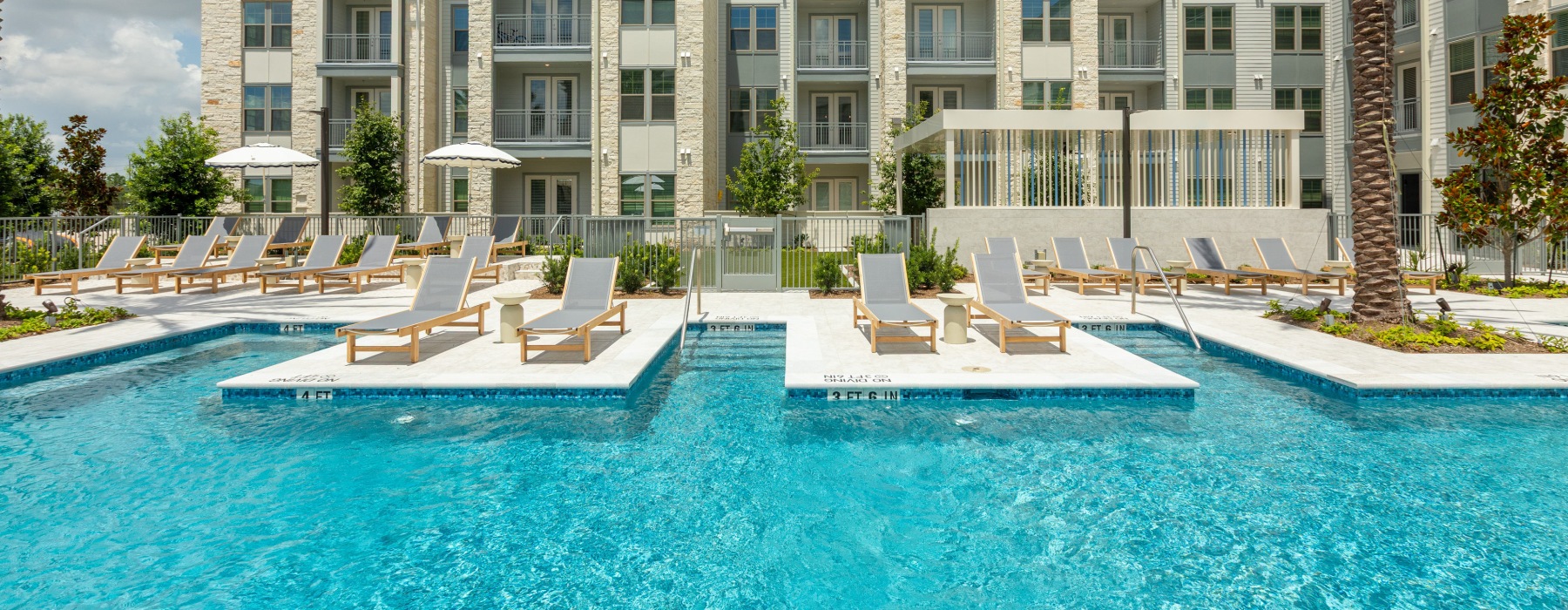 A modern residential complex with two swimming pools, lounge chairs, and a backdrop of a multi-story building under a blue sky.