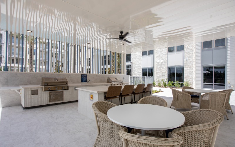 Outdoor grill area featuring a stainless steel grill, marble countertop, bar stools, and a white dining table with woven chairs in a bright, modern setting.