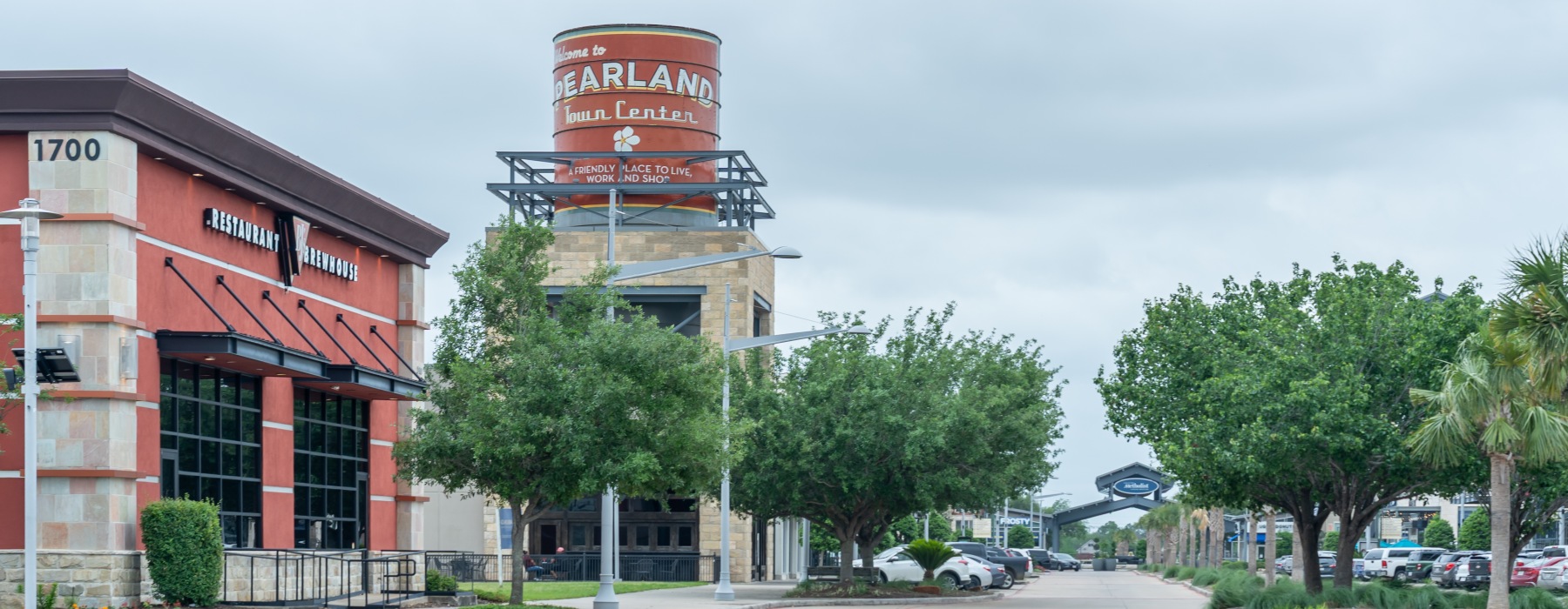 A shopping center features a large red cylindrical water tower labeled "Welcome to Pearland Town Center." To the left, a building with the name "Restaurant Greenhouse" is visible. The sky is cloudy, and trees line the paved pathway with parked cars nearby