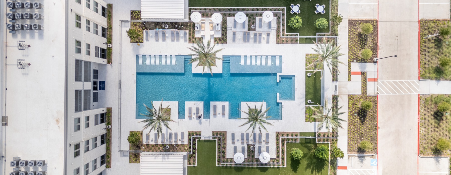 Aerial view of a modern apartment complex with a central swimming pool surrounded by lounge chairs, palm trees, and landscaped gardens, alongside a paved path and parking area.